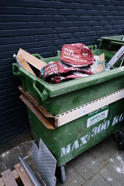 A large green wheeled rubbish skip filled with red and black bags containing building materials or waste, placed outdoors against a dark blue brick wall. The bags are marked with product labels, and a section of a white plastic barrier with green and black text is visible on the side of the skip. The scene is set on a concrete or paved surface, with a small drain cover nearby, and the overall environment suggests an outdoor area prepared for rubbish removal or waste collection. Details such as the texture of the waste bags, the sturdy plastic finish of the skip, and the contrasting dark brick background highlight a typical site for private waste disposal or on-site clearance managed by a waste removal service like Rubbish Removal Fulham.