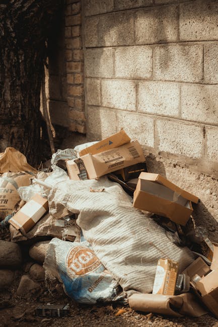 A large green wheeled rubbish skip filled with red and black bags containing building materials or waste, placed outdoors against a dark blue brick wall. The bags are marked with product labels, and a section of a white plastic barrier with green and black text is visible on the side of the skip. The scene is set on a concrete or paved surface, with a small drain cover nearby, and the overall environment suggests an outdoor area prepared for rubbish removal or waste collection. Details such as the texture of the waste bags, the sturdy plastic finish of the skip, and the contrasting dark brick background highlight a typical site for private waste disposal or on-site clearance managed by a waste removal service like Rubbish Removal Fulham.