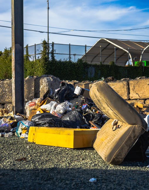 A pile of mixed waste materials and rubbish bags situated on a rough asphalt surface outside, with a weathered, discarded beige tire leaning heavily against the pile. The debris includes crumpled black, white, and transparent plastic bags filled with waste, some of which are torn open, revealing discarded packaging and miscellaneous refuse. A bright yellow plastic container lies open among the rubbish, partially covered by waste, suggesting collection or temporary storage. In the background, a low stone wall made up of irregularly shaped, rough-hewn stones separates the rubbish from an area with green shrubbery, a tall metal utility pole, and overhead power lines. Behind the fence, a large, semi-circular, white fabric-covered sports or storage structure, supported by metal framework, can be seen under a clear blue sky with scattered clouds, indicating the scene is during daytime. The environment conveys an outdoor setting requiring waste removal, reflecting the importance of on-site clearance or private disposal services that Rubbish Removal Fulham provides, especially when dealing with general rubbish accumulation outside residential or commercial premises.