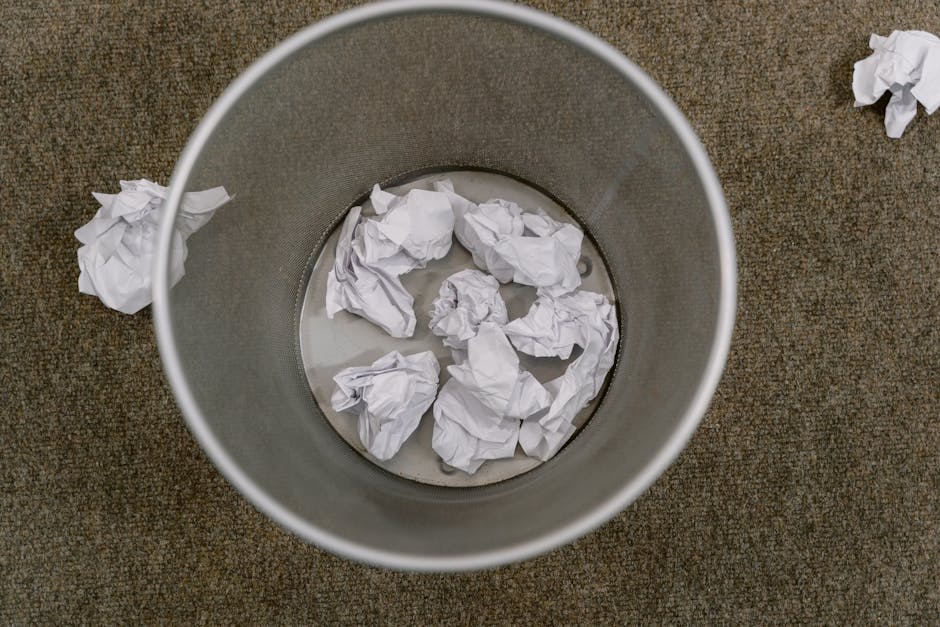 A pile of mixed waste materials and rubbish bags situated on a rough asphalt surface outside, with a weathered, discarded beige tire leaning heavily against the pile. The debris includes crumpled black, white, and transparent plastic bags filled with waste, some of which are torn open, revealing discarded packaging and miscellaneous refuse. A bright yellow plastic container lies open among the rubbish, partially covered by waste, suggesting collection or temporary storage. In the background, a low stone wall made up of irregularly shaped, rough-hewn stones separates the rubbish from an area with green shrubbery, a tall metal utility pole, and overhead power lines. Behind the fence, a large, semi-circular, white fabric-covered sports or storage structure, supported by metal framework, can be seen under a clear blue sky with scattered clouds, indicating the scene is during daytime. The environment conveys an outdoor setting requiring waste removal, reflecting the importance of on-site clearance or private disposal services that Rubbish Removal Fulham provides, especially when dealing with general rubbish accumulation outside residential or commercial premises.