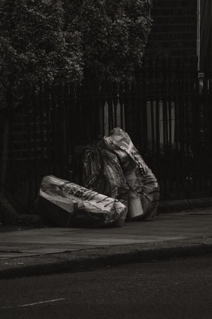 Three large black plastic rubbish bags, some with visible white and black printed text, are stacked against a black metal fence on a pavement. The bags appear filled with waste material, with creases and crumpled textures on their surfaces. Behind the fence, portions of lush green foliage from trees or bushes are visible, providing a natural backdrop. To the right, part of a dark building structure can be seen, with a slight glow suggesting ambient outdoor lighting. The scene suggests an informal rubbish collection point on a street, which may relate to private waste handling services like those offered by Rubbish Removal Fulham, providing an alternative to traditional council rubbish collection for disposing of household or garden waste safely and efficiently. The overall scene emphasizes the practical aspect of rubbish disposal outside residential or urban areas, highlighting the importance of managed waste removal in urban environments.