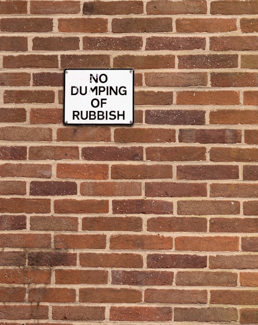 A square white metal sign mounted on a red brick wall, displaying the message 'NO DUMPING OF RUBBISH' in black capital letters, with the word 'DUMPING' partially obscured by a black vertical line. The brick wall features a horizontal pattern of reddish-brown bricks with varying shades and textures, some darker and more weathered, laid in a staggered brickbond pattern with light-colored mortar joints. The sign is positioned slightly above the center of the image, and the overall scene suggests a restricted area for rubbish disposal. The wall appears to be part of an outdoor setting, possibly near a rubbish removal or waste collection point, consistent with the services offered by Rubbish Removal Fulham for private waste handling or alternative disposal options.