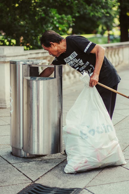 A middle-aged man with dark hair, wearing a black T-shirt with white text and dark trousers, is seen standing outdoors on a paved walkway during daytime. He is using a wooden stick or pick to lift the lid of a stainless steel public rubbish bin, which has a rounded rectangular shape and a shiny, smooth surface. The trash bin is positioned against a low stone wall with a decorative railing, and behind it, there are lush green trees and foliage, indicating a park or garden setting. The man is holding a large white plastic rubbish bag filled with waste, partially resting on the ground, and appears to be in the process of discarding or collecting rubbish. The scene is well-lit with natural daylight, creating a clear and detailed view of the person's activity focused on waste disposal or collection, consistent with the context of rubbish removal services.