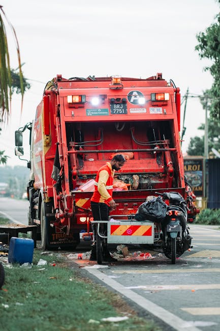 A worker dressed in a high-visibility yellow and orange vest is standing next to a large, bright red commercial rubbish collection truck on the side of a paved street. The truck's rear compartment is open, revealing mechanical components and a collection of black rubbish bags and debris inside. The worker appears to be loading or inspecting waste, with a small metal barrier or guard placed on the pavement nearby. The scene is outdoors with trees and utility poles visible in the background, and the environment suggests an urban or suburban area where private waste collection services, such as Rubbish Removal Fulham, operate. The overcast sky and the surrounding greenery provide a neutral atmosphere, emphasizing the focus on waste collection processes and vehicle readiness for rubbish removal tasks.