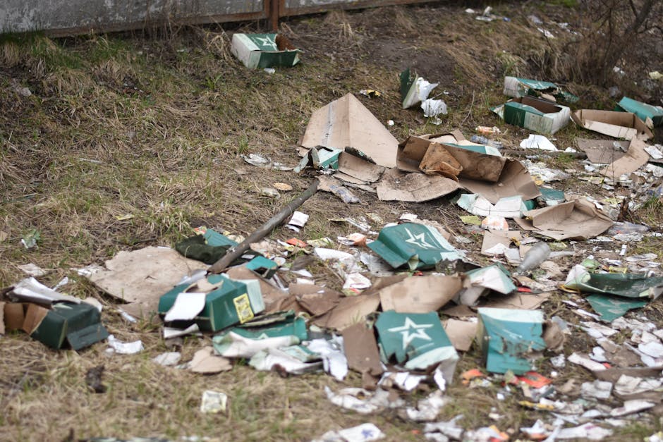 A large outdoor scene displaying scattered cardboard packaging, flattened and torn, with some boxes displaying a white star logo on a green background, lying among dry grass and dirt on a slope. The debris includes broken boxes, shredded paper, plastic bottles, and miscellaneous litter, with a metal pipe partly visible among the rubbish. In the background, there is a weathered brick or metal fence at the top of the slope. The environment appears neglected, highlighting the need for professional waste collection services such as those provided by Rubbish Removal Fulham. The scene reflects a typical example of event-related waste or litter left after a gathering, demonstrating the importance of on-site clearance and private waste handling for effective rubbish removal during large events or clean-up operations.