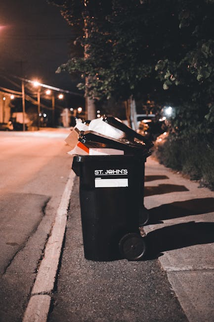 A black wheeled rubbish bin positioned on a sidewalk beside a curb on a quiet street at night. The bin has a white label with the text 'St. John's' and is filled with various waste items, including flattened cardboard boxes, plastic packaging, and other miscellaneous refuse. The street is illuminated by streetlights creating warm, orange-toned lighting, and the background shows blurred trees, parked cars, and buildings faintly visible under the low light. The scene suggests a typical collection point for private or independent waste disposal, reflecting an alternative to regular council rubbish collection. The bin's appearance, along with the urban environment, aligns with household or commercial rubbish removal services, such as those offered by Rubbish Removal Fulham, who provide on-site clearance and private waste handling options for residents and businesses in Fulham.